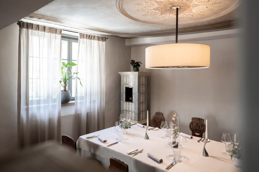 Tavola apparecchiata al ristorante Löwengrube con sedie antiche in legno, pianta alla finestra, lampada di design e stufa in maiolica. ©Löwengrube/Alex Filz