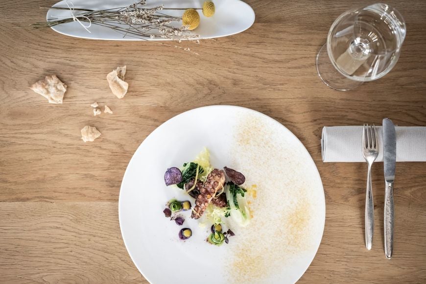 Piatto di pesce su tavolo in legno con bicchiere di vino bianco, Schüttelbrot, posate e decorazione floreale. ©Löwengrube/Alex Filz